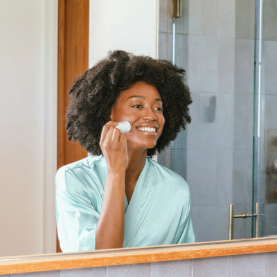 Woman in a robe applying Refreshing Water Hydration Stick sunscreen while smiling in front of a bathroom mirror.
