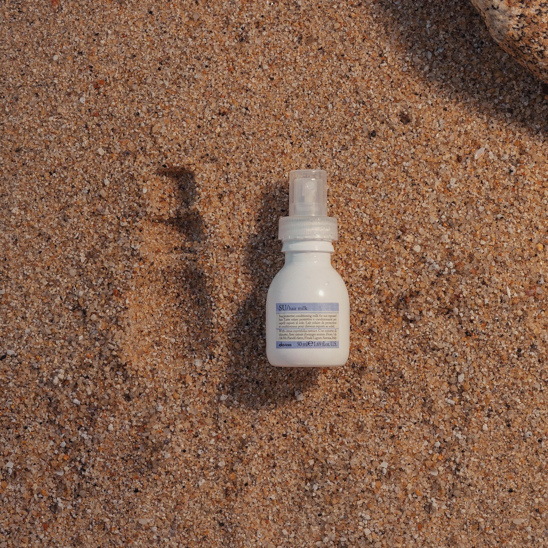 A travel size Davines SU Hair Milk lays in the sand next to an imprint of the bottle in the sand.