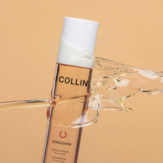 A bottle of Gm Collin Sensiderm Cleansing Oil is suspended against a peach tone background, being splashed with oil. 