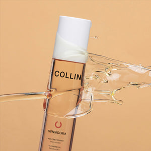 A bottle of Gm Collin Sensiderm Cleansing Oil is suspended against a peach tone background, being splashed with oil. 