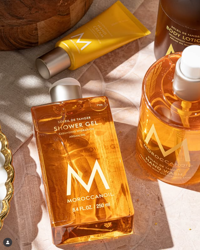 Bottles of Moroccanoil Soleil De Tanger scented shower gel, hand wash, body lotion,  and hand cream are sunlit, against a stone background next to a wooden basket.