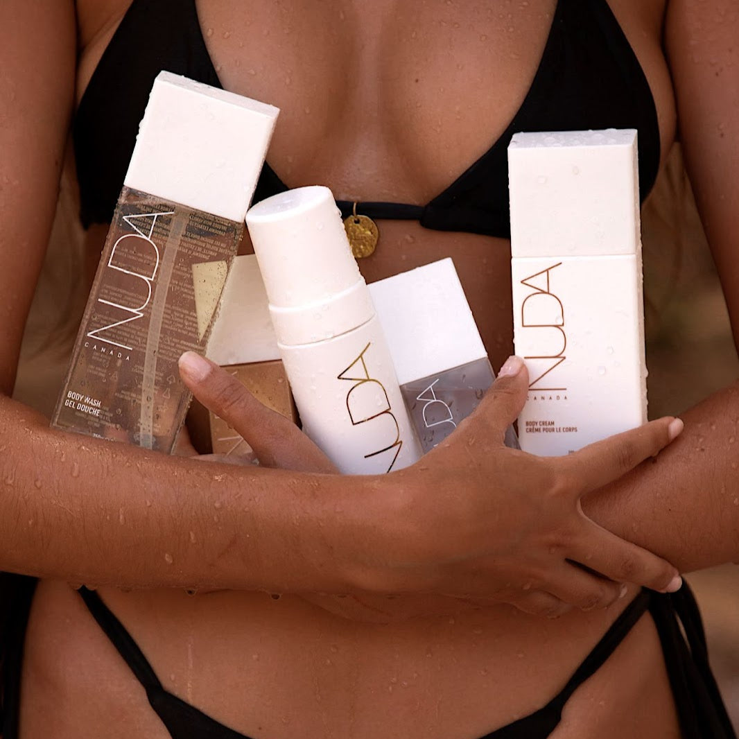A tanned woman in a bikini, dripping water, holds various Nuda product bottles including Body Wash, Body Cream and Face Tan Water, in her arms. 