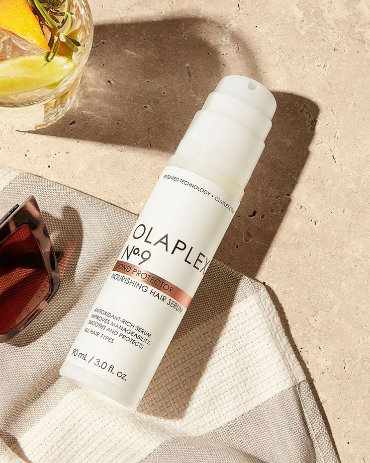 Olaplex N9 Bond Protector bottle lays on a stone background next to a cocktail and a pair of sunglasss. 