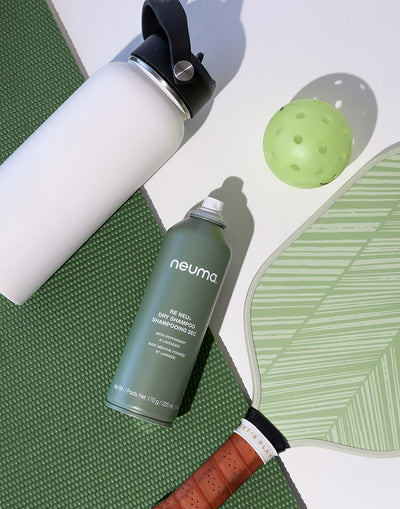 Neuma Re Neu Dry Shampoo bottle next to a white water bottle and a green paddleball on a textured green surface.