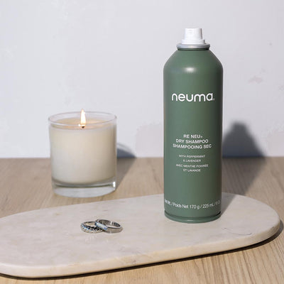 Neuma Re Neu Dry Shampoo canister on a marble tray with a lit candle and rings, showcasing a fresh and stylish bathroom setting.
