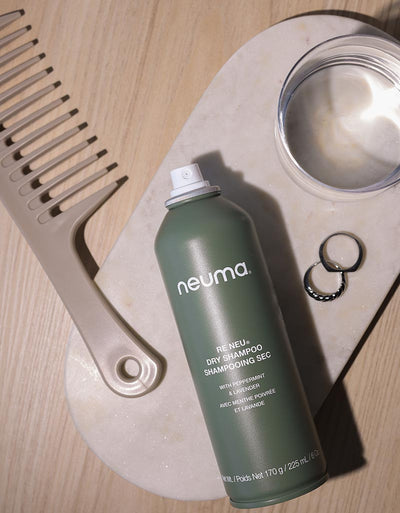 Neuma Re Neu Dry Shampoo can on marble tray with comb, rings, and glass of water, showcasing hair revitalizing product.