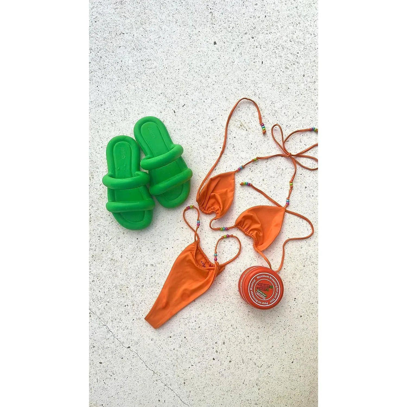 Bright orange bikini set paired with green slide sandals and a carrot cream tanning product on a light surface.