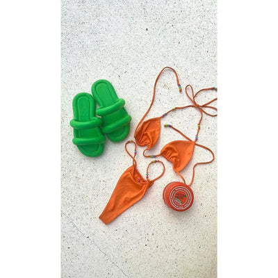 Bright orange bikini set paired with green slide sandals and a carrot cream tanning product on a light surface.