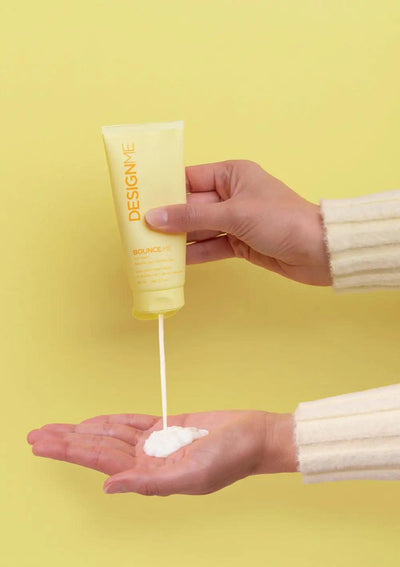 Hand pouring BOUNCE.ME curl balm into palm against a bright yellow background.