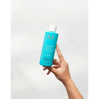 Hand holding Moroccanoil Extra Volume Shampoo bottle against a neutral background.