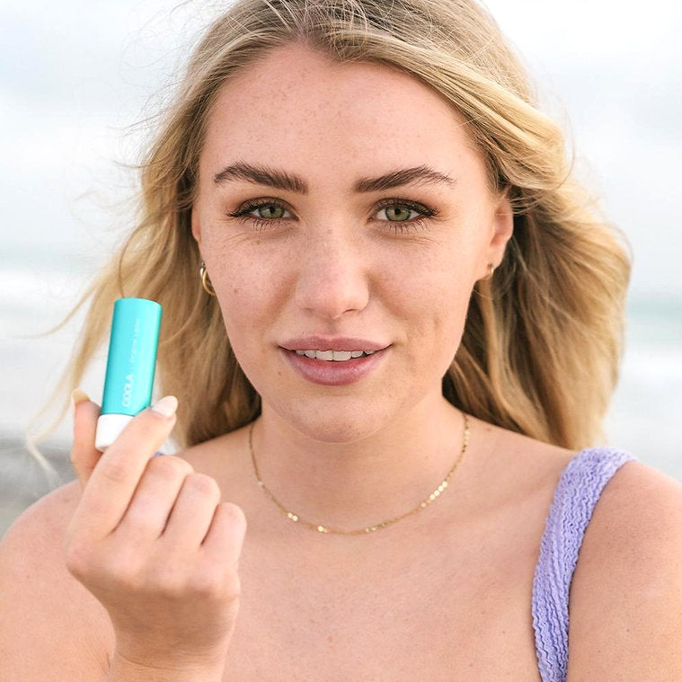 Woman holding Classic Liplux® Organic Lip Balm SPF 30, showcasing radiant skin and beachy backdrop.