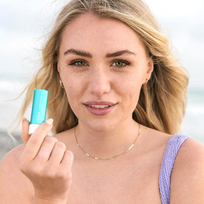 Woman holding Classic Liplux® Organic Lip Balm SPF 30, showcasing radiant skin and beachy backdrop.