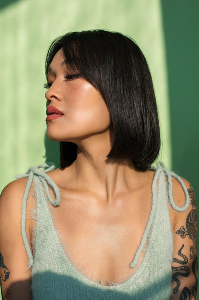Woman with sleek black bob hairstyle and tattoos posing against a green background, showcasing natural beauty.