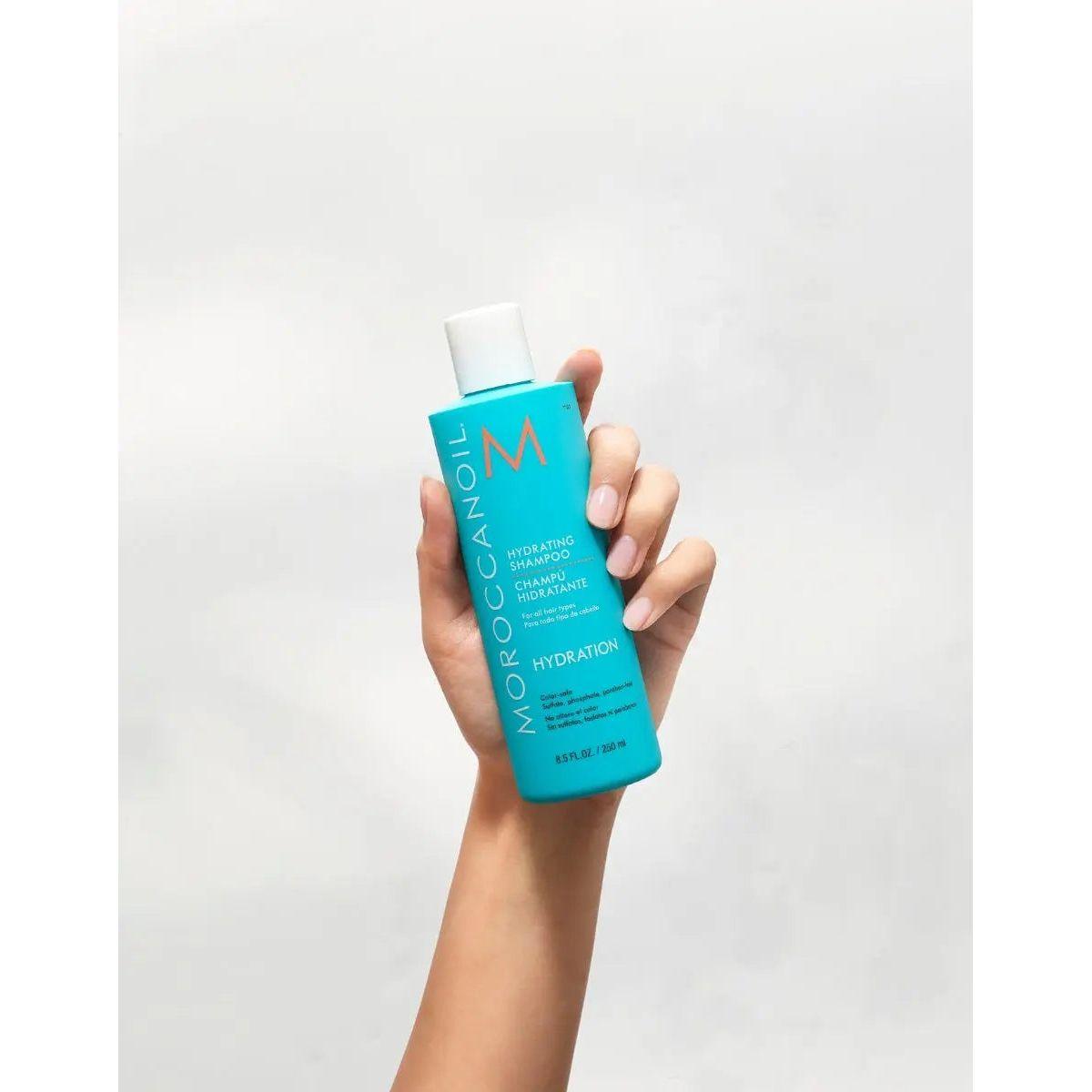 Hand holding Moroccanoil Hydrating Shampoo bottle, showcasing its vibrant blue packaging for healthy hair care.