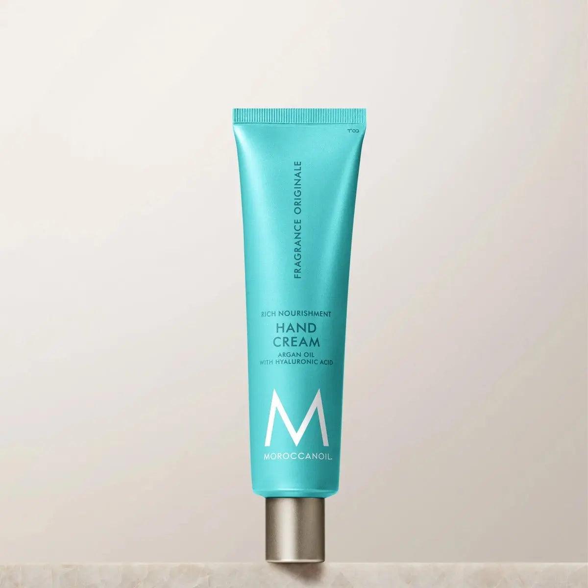 Moroccanoil Hand Cream featuring argan oil and hyaluronic acid in a turquoise tube on a neutral background.
