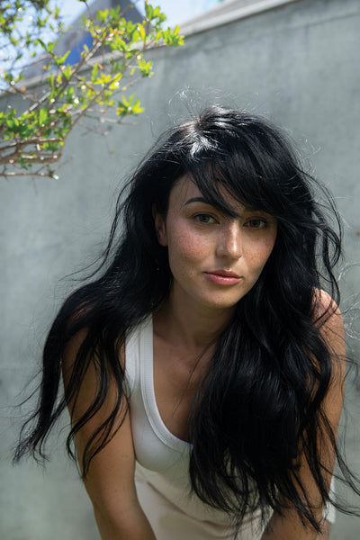 Woman with long, wavy black hair wearing a white tank top, showcasing natural beauty in a sunny outdoor setting.