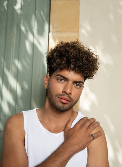 Young man with curly hair posing casually against a textured green wall, showcasing a stylish look.