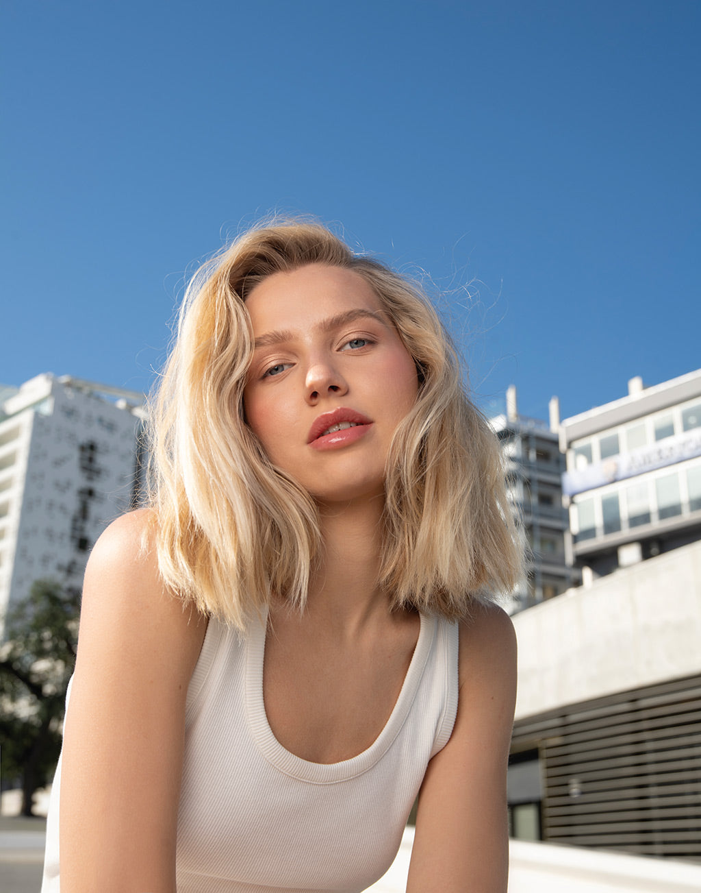 Young woman with blonde hair and natural makeup, smiling outdoors against a city backdrop. Sunlit, casual summer vibe.
