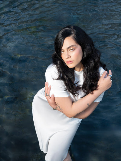 Model with wavy hair poses gracefully in water, showcasing a serene and stylish look in a white dress.
