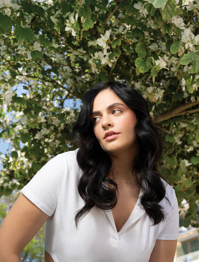 Woman with wavy black hair in white top, posing in front of blooming tree, showcasing healthy hair and natural beauty.