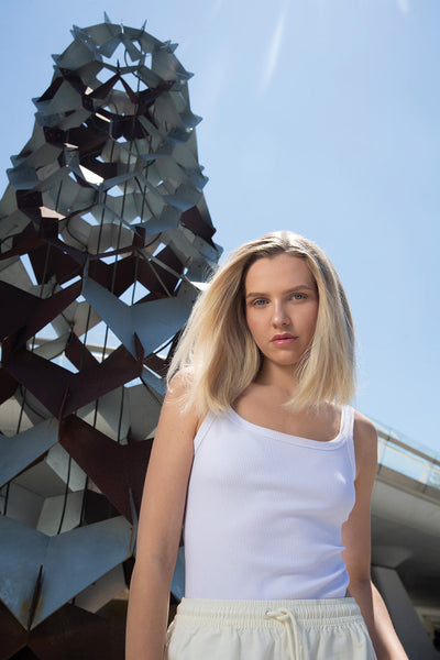 Young woman in a white tank top stands in front of a modern sculpture, showcasing her long blonde hair in bright sunlight.