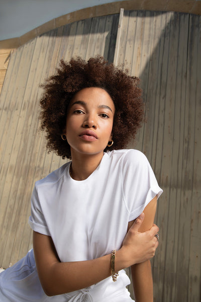 Model with curly hair wearing a white shirt, posing against a wooden background in natural light.