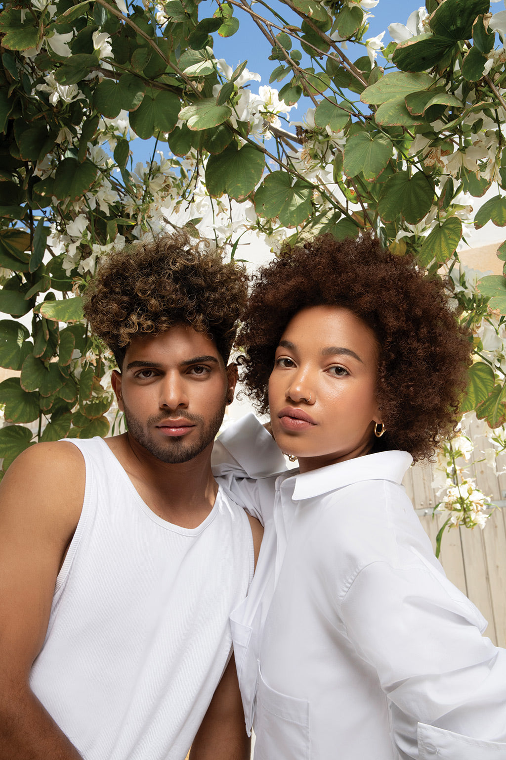 Two models with curly hairstyles posing outdoors among blooming flowers, showcasing natural curls and radiant skin.