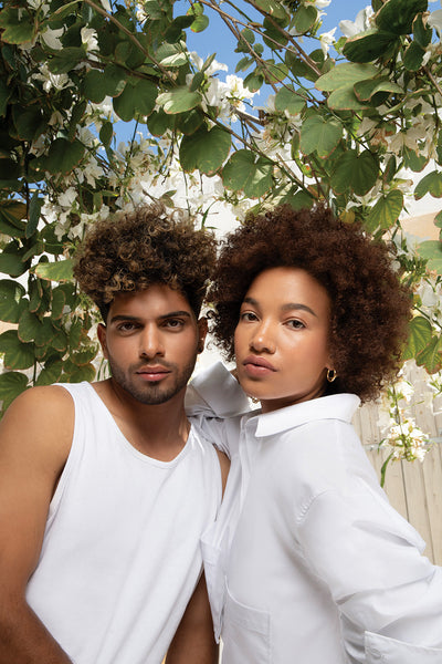 Two models with curly hairstyles posing outdoors among blooming flowers, showcasing natural curls and radiant skin.