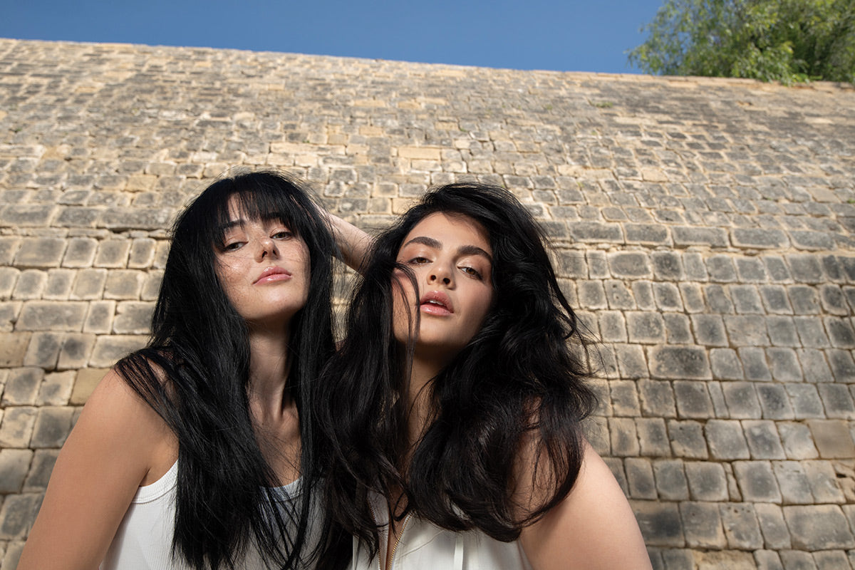 Two women with long, flowing hair outdoors, showcasing healthy, smooth hair against a stone wall backdrop.