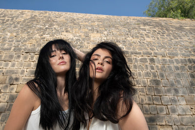 Two women with long, flowing hair outdoors, showcasing healthy, smooth hair against a stone wall backdrop.