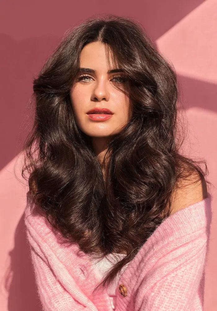 Young woman with voluminous, wavy hair in a pink sweater against a pink background, showcasing healthy, glossy locks.