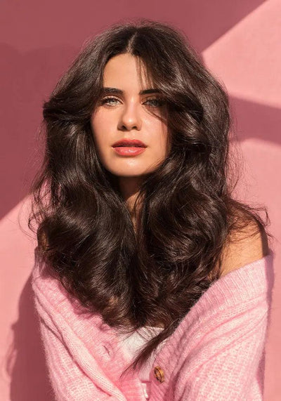 Young woman with voluminous, wavy hair in a pink sweater against a pink background, showcasing healthy, glossy locks.