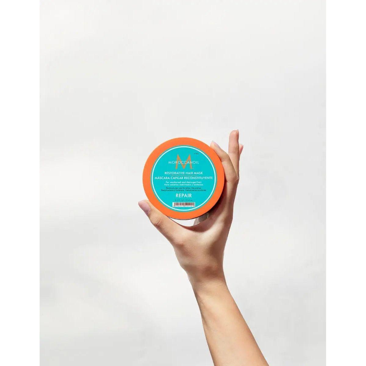 Hand holding a bright orange jar of Restorative Repair Hair Mask for deep hair repair and hydration.