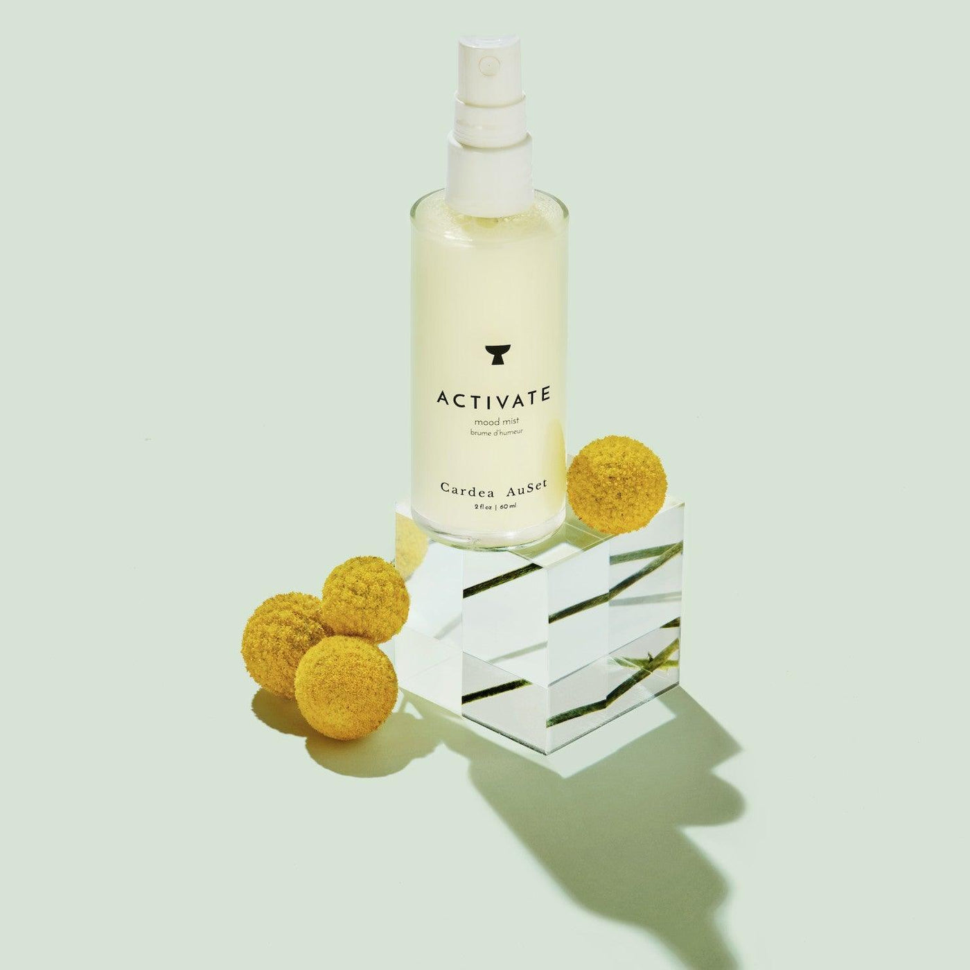 Activate facial mist by Cardea AuSet on a geometric glass block with yellow flowers, green background, skincare product.