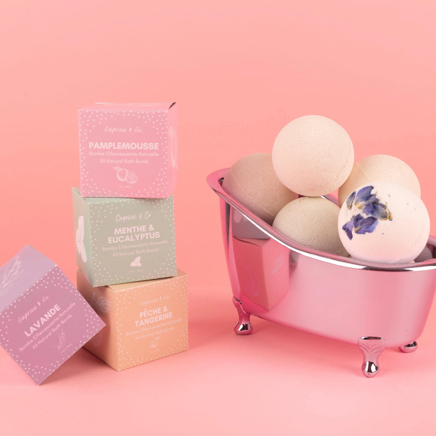 All natural bath bombs in a silver tub with colorful packaging on a pink background.