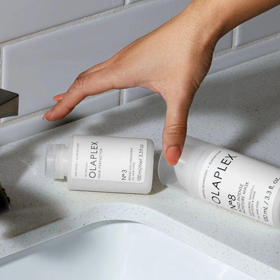 Hand reaching for Olaplex Nº.3 Hair Perfector and Nº.8 Bond Intense Moisture Mask on a bathroom countertop.