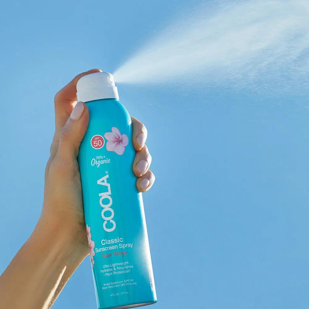 Hand spraying Classic Body Organic Sunscreen Spray SPF 50 against a clear blue sky for sun protection.