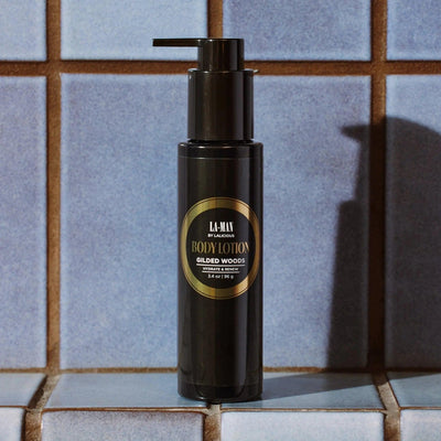 Gilded Woods Body Lotion in a sleek pump bottle against blue tiled background, offering hydration and nourishment for skin.