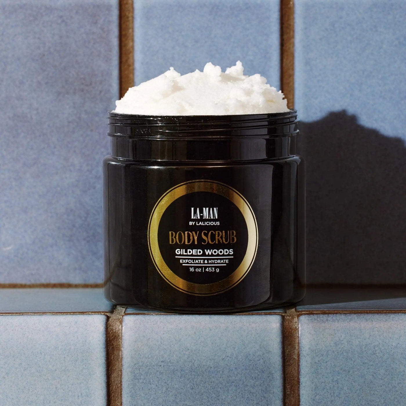 Gilded Woods Body Scrub jar on a tiled surface, featuring a rich, creamy exfoliating scrub.