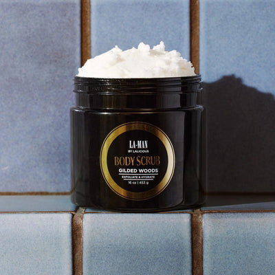 Gilded Woods Body Scrub jar on a tiled surface, featuring a rich, creamy exfoliating scrub.