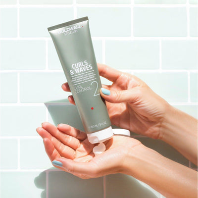 Hand holding Goldwell Curls & Waves Curl Control Moisturizing Curl Cream in a stylish bathroom setting.