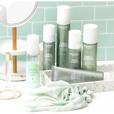 Goldwell Curls & Waves curl care products displayed on a bathroom counter with a mirror and decorative accessories.