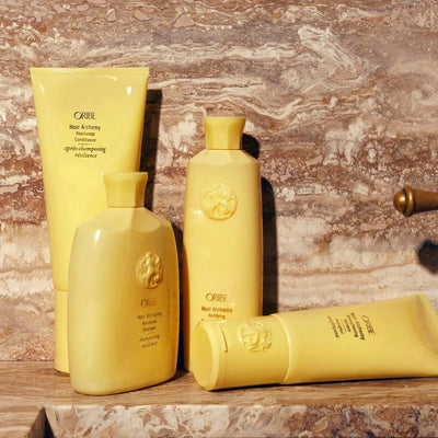 Oribe Hair Alchemy shampoo and conditioner bottles in yellow packaging displayed on a marble countertop.