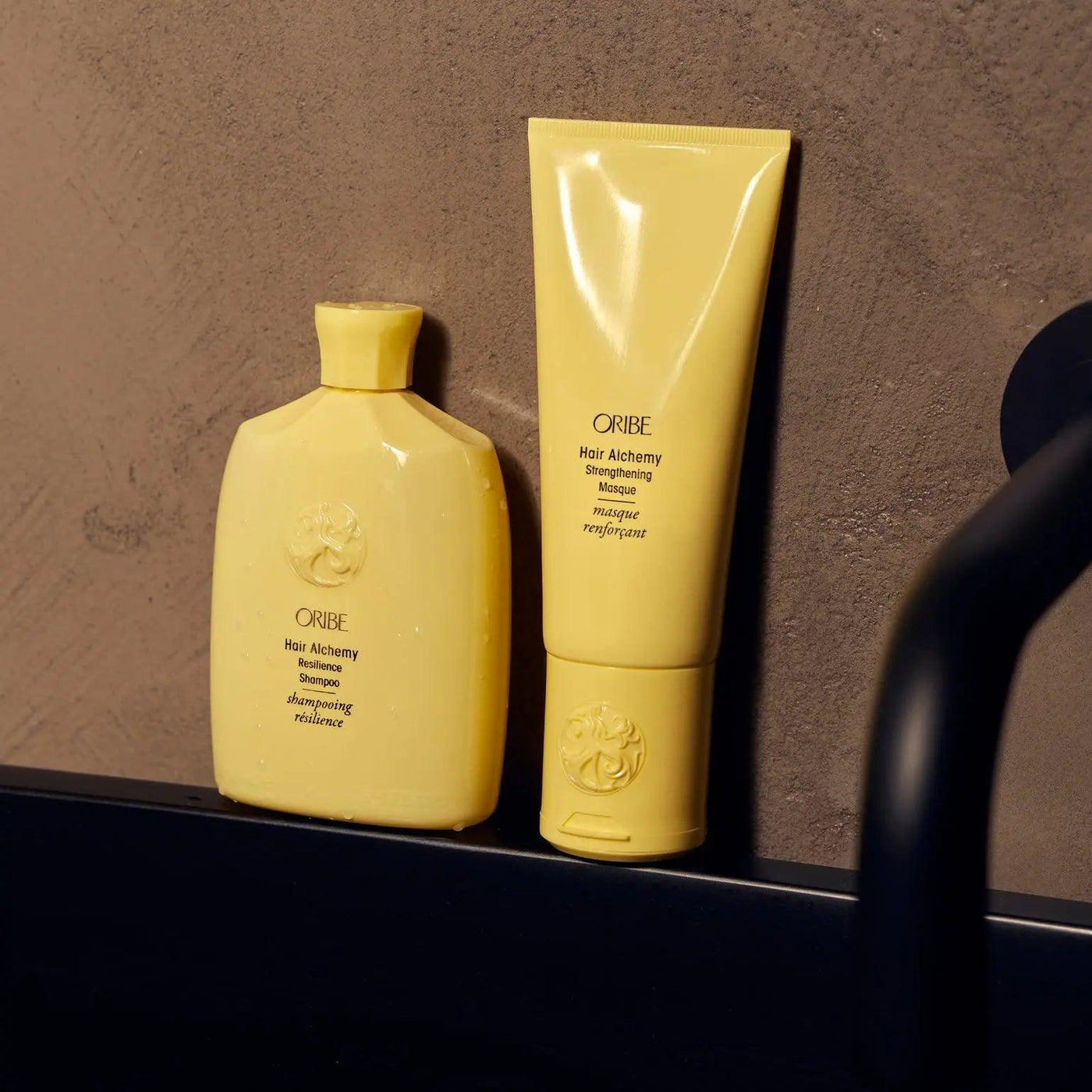 Two yellow tubes of Oribe Hair Alchemy treatment products on a bathroom countertop against a textured wall.