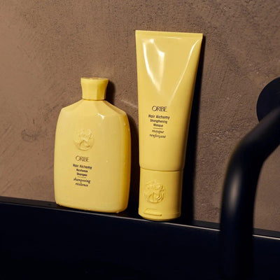 Two yellow tubes of Oribe Hair Alchemy treatment products on a bathroom countertop against a textured wall.