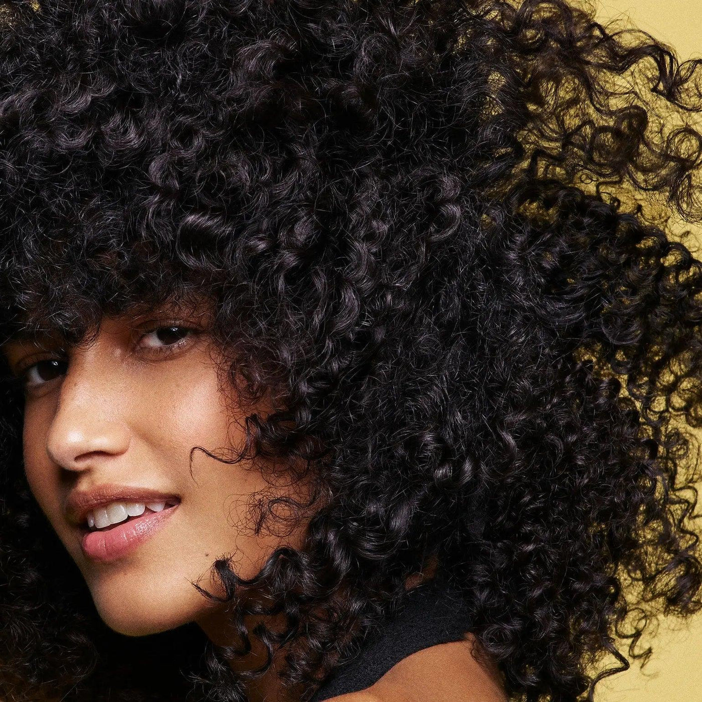 Close-up of a woman with voluminous curly hair, showcasing healthy, defined curls against a bright background.