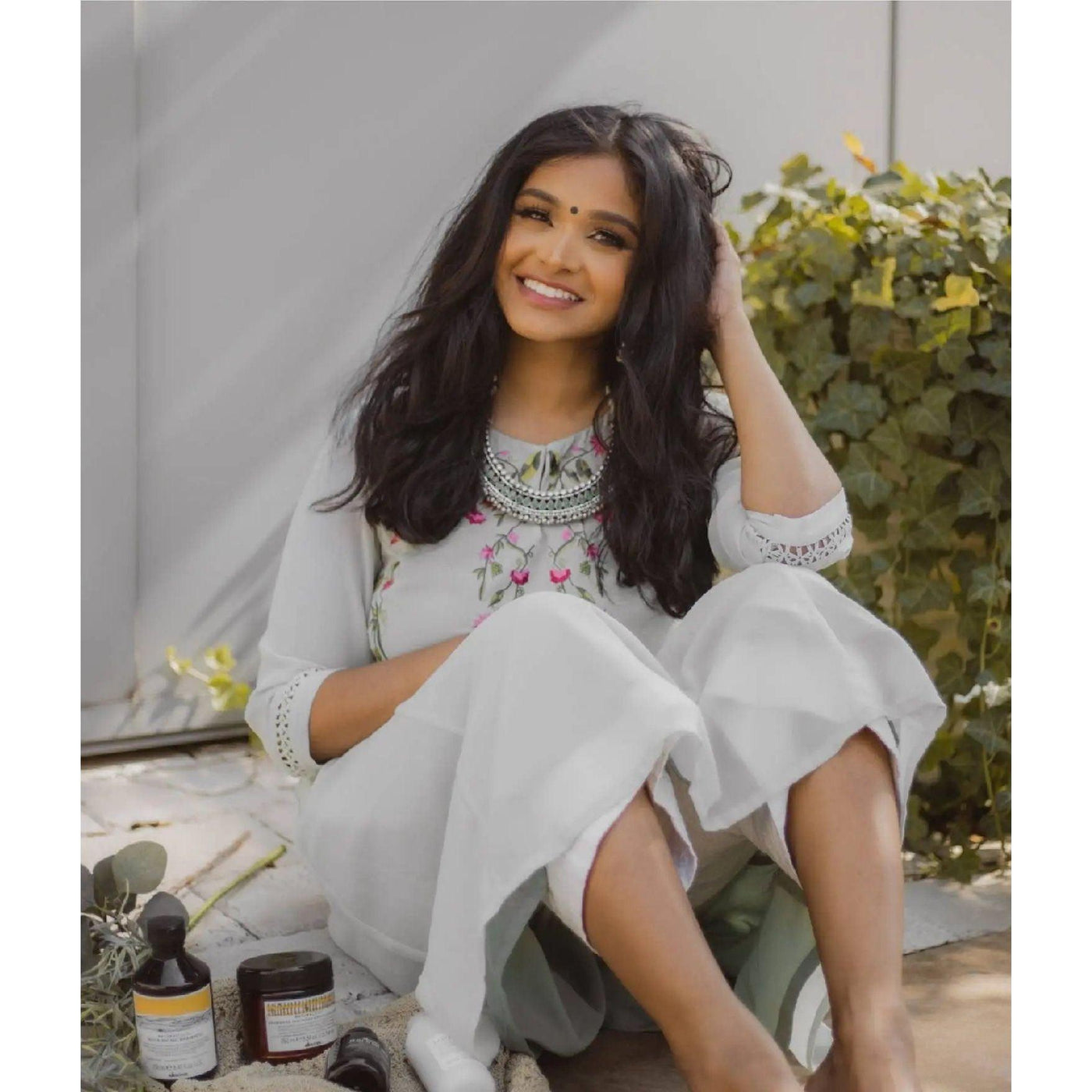 Smiling woman in traditional attire with natural waves sitting among hair care products in a sunny setting.