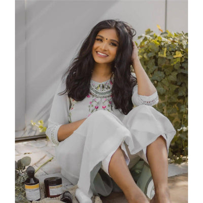 Smiling woman in traditional attire with natural waves sitting among hair care products in a sunny setting.