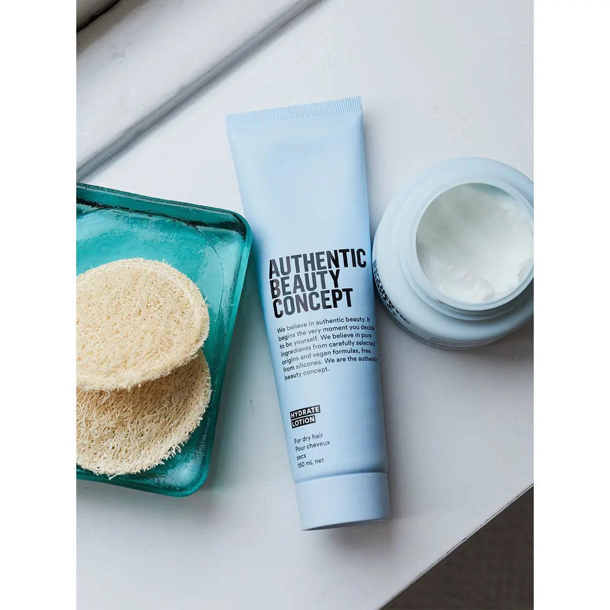 Authentic Beauty Concept Hydrate Lotion tube with natural loofah sponges, promoting shine and protection for dry hair.