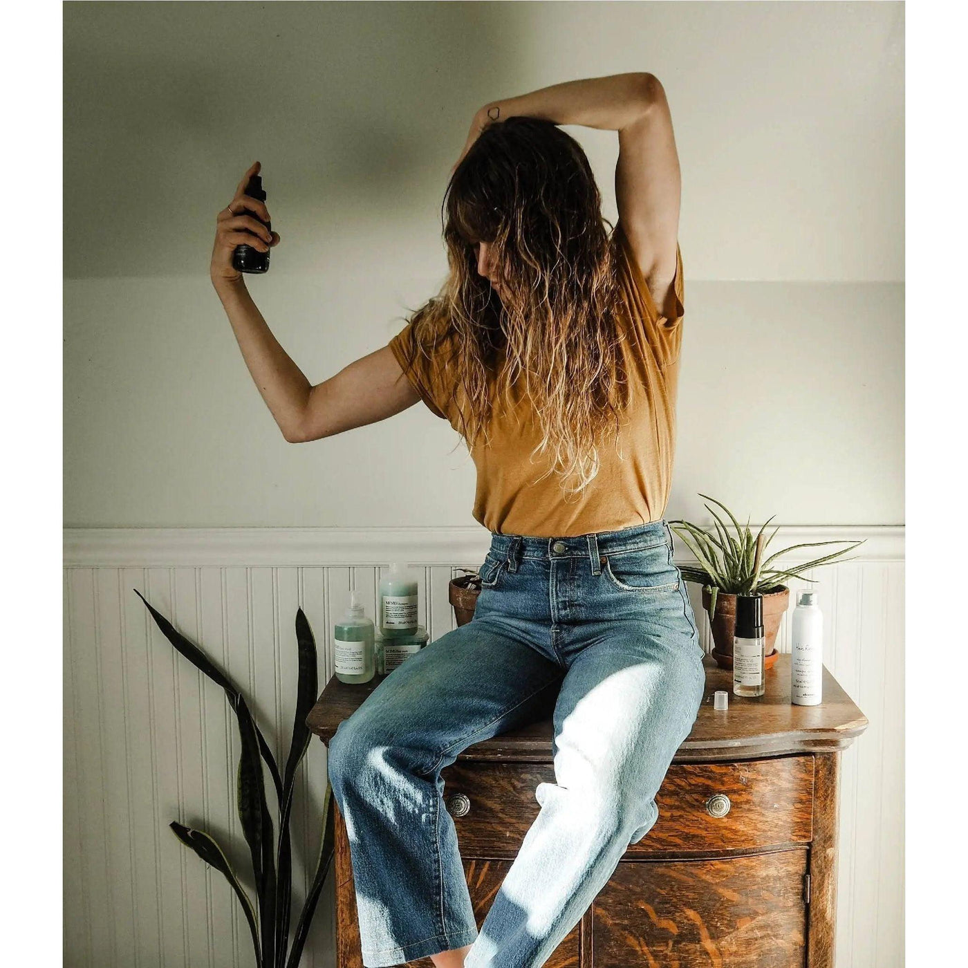 Woman using a hair styling spray while sitting on a wooden dresser, showcasing a low-maintenance textured hair routine.